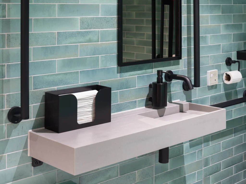 An image of a teal tiled wall in a DDA DocM accessible washroom. The focus is on the basin area, with other accessories clustered around handwashing also on view. 