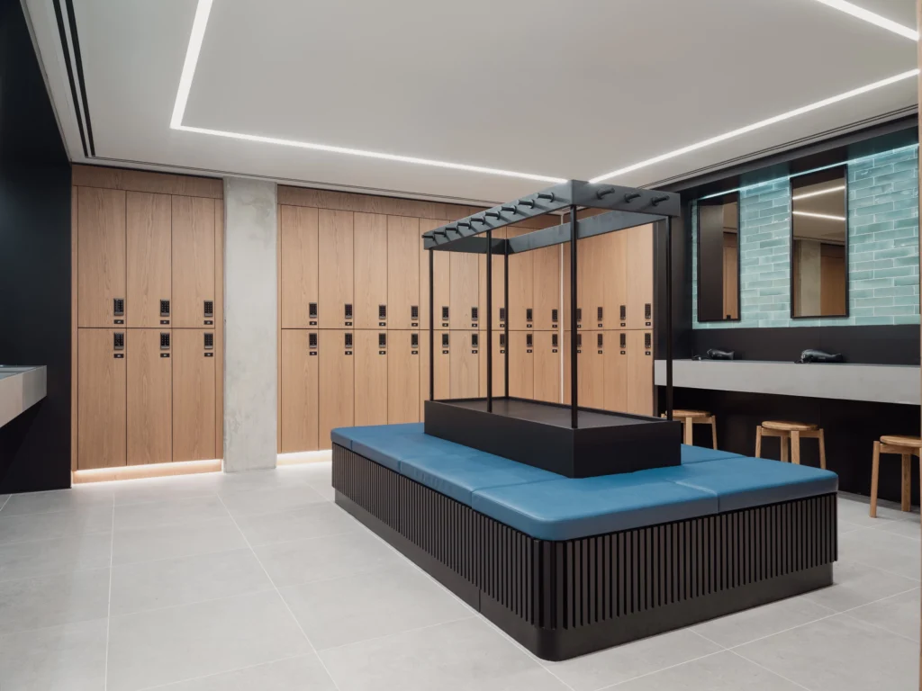 A changing room in teal tile and neutral accents, with a central bench and lockers, basins, and vanities around the edges of the room. 